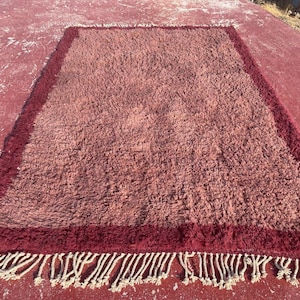 May include: A rectangular area rug featuring a shaggy, textured surface in a muted mauve tone. The rug is framed by a darker, deep burgundy border and finished with long, cream-colored fringe tassels along the bottom edge of the piece.
