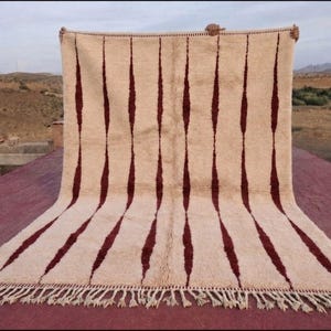 Peut inclure: Un grand tapis rectangulaire au fond crème orné de rayures verticales en forme de losanges marron foncé s'étendant de haut en bas. Le tapis présente un poil épais et texturé, terminé par de longues franges nouées sur le bord inférieur.