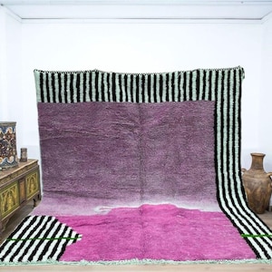 May include: A large, rectangular rug with a gradient of colors, from deep purple to bright pink. The rug has a black and white striped border. The rug is displayed against a white wall.