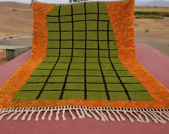 May include: A rectangular rug with a green grid pattern on a green background, framed by an orange border. The rug has a fringe of white tassels along one edge. The rug is displayed outdoors.
