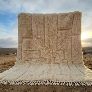 May include: A large, rectangular, off-white area rug featuring a thick, high-pile shaggy texture. The surface displays a subtle, raised geometric pattern created by varying pile heights, finished with a fringe of knotted tassels along the bottom edge.