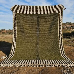 May include: Olive green rectangular rug with a textured surface. The rug has a border of alternating black and white stripes, and white fringe along the bottom edge. The rug is displayed outdoors against a backdrop of a field and blue sky.