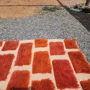 May include: A rectangular rug with a cream-colored background and a pattern of large, shaggy, rectangular shapes in shades of orange and red. The rug is placed on a gravel surface, with a glimpse of dirt and vegetation in the background.