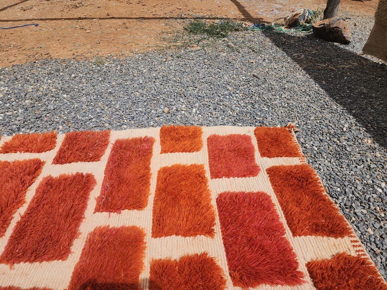 May include: A rectangular rug with a cream-colored background and a pattern of large, shaggy, rectangular shapes in shades of orange and red. The rug is placed on a gravel surface, with a glimpse of dirt and vegetation in the background.