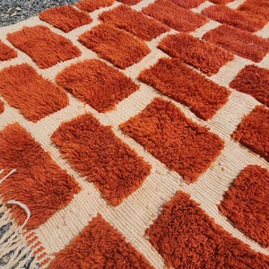 May include: A textured rug with a repeating pattern of orange rectangular tufts on a cream-colored background. The rug has a fringe on one edge. The rug is made of a soft, plush material.