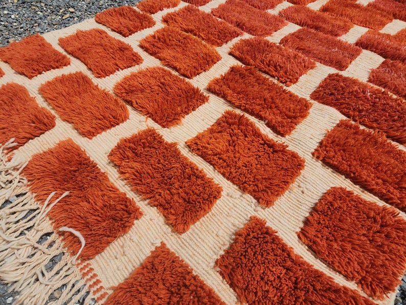 May include: A textured rug with a repeating pattern of orange rectangular tufts on a cream-colored background. The rug has a fringe on one edge. The rug is made of a soft, plush material.