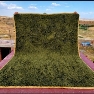May include: A rectangular, deep olive green shaggy blanket featuring a thick, mustard yellow crocheted border along all four edges. The blanket has a dense, textured pile surface and is held up to display its full size and uniform color.