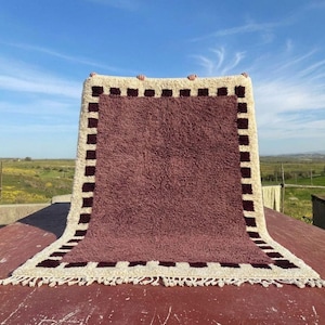 May include: A rectangular rug with a burgundy center and a cream-colored border. The border features a repeating pattern of dark burgundy squares. The rug is displayed outdoors against a blue sky.