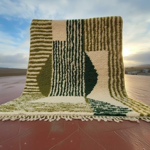 May include: A rectangular rug with a geometric pattern in shades of green, beige, and white. The rug has a textured, tufted design with a fringe along the bottom edge. The rug is displayed outdoors, with a view of the sky and landscape in the background.