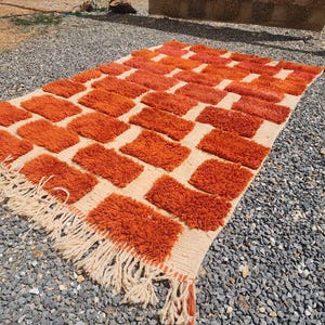 May include: An orange and cream-colored rug with a geometric pattern of rectangular shapes. The rug has a shaggy texture and a fringe along the edges. The rug is made of a soft material and is suitable for a living room or bedroom.