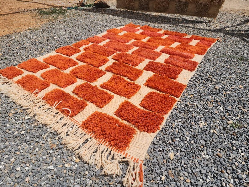 May include: An orange and cream-colored rug with a geometric pattern of rectangular shapes. The rug has a shaggy texture and a fringe along the edges. The rug is made of a soft material and is suitable for a living room or bedroom.