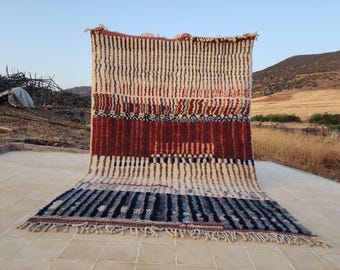May include: A rectangular rug with a striped pattern in cream, red, and navy blue. The rug has a textured appearance with a fringe along the bottom edge. The background shows a natural outdoor setting with a clear sky and hills.