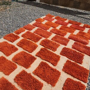 May include: A rectangular rug with a cream-colored background and a pattern of orange rectangular blocks. The blocks have a shaggy texture, and the rug is placed on a gravel surface.