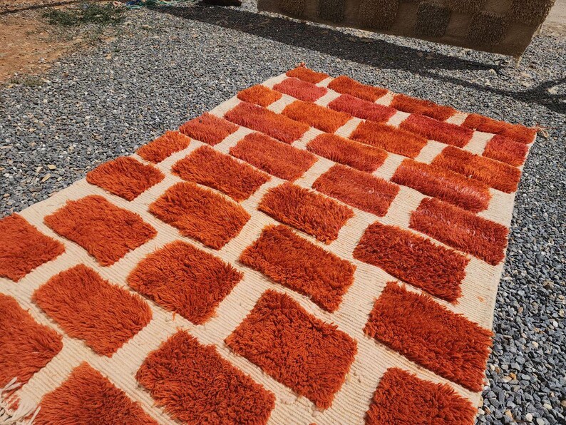 May include: A rectangular rug with a cream-colored background and a pattern of orange rectangular blocks. The blocks have a shaggy texture, and the rug is placed on a gravel surface.