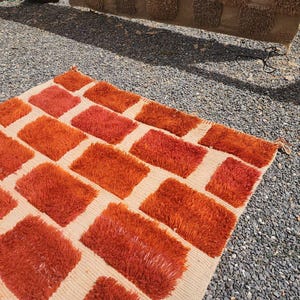 May include: A rectangular rug with a cream-colored background and a pattern of orange rectangular blocks. The blocks have a shaggy texture, and the rug is placed on a gravel surface.