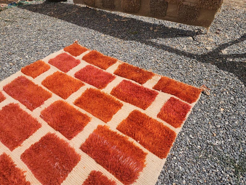 May include: A rectangular rug with a cream-colored background and a pattern of orange rectangular blocks. The blocks have a shaggy texture, and the rug is placed on a gravel surface.