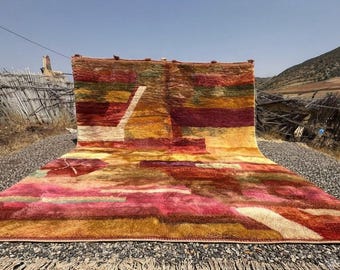 May include: A large rectangular area rug featuring a vibrant, abstract color palette of deep red, burnt orange, mustard yellow, olive green, and magenta. The rug displays a thick, textured pile with geometric shapes and a fringed edge.