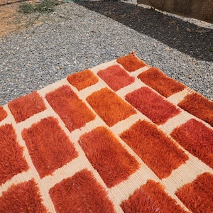 May include: A rectangular rug with a repeating pattern of orange rectangles on a beige background. The rug has a shaggy texture, and the orange rectangles vary in shade, creating visual interest. The rug is placed outdoors on a surface of gravel.