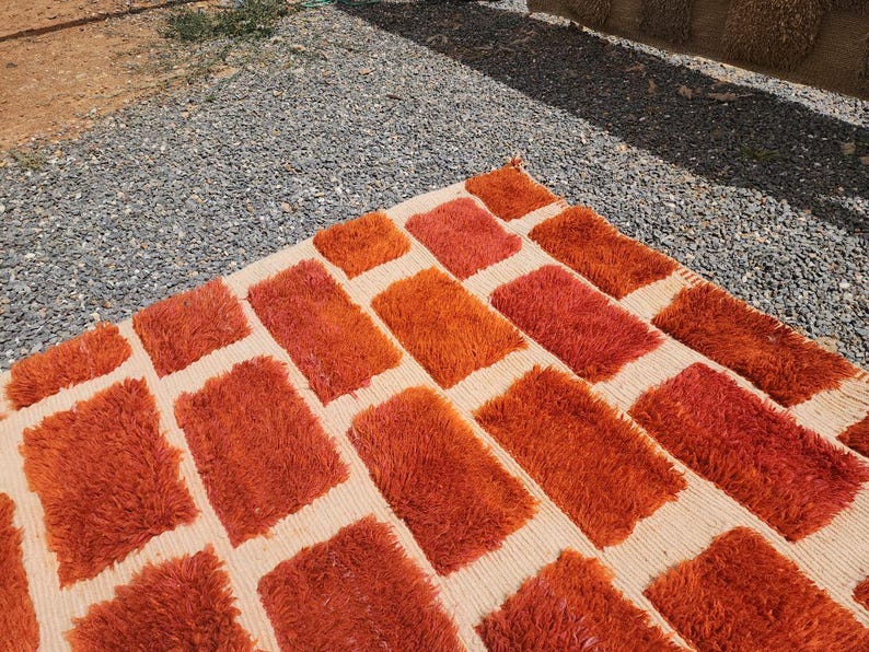 May include: A rectangular rug with a repeating pattern of orange rectangles on a beige background. The rug has a shaggy texture, and the orange rectangles vary in shade, creating visual interest. The rug is placed outdoors on a surface of gravel.