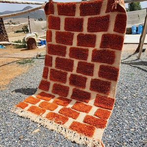 May include: A rectangular rug with a brick-like pattern in shades of orange and beige. The rug is hanging outdoors, with a fringe along the bottom edge. The background shows other textiles and a rural landscape.