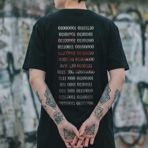 Könnte beinhalten: Ein schwarzes Kurzarm-T-Shirt mit einer vertikalen Spalte aus Binärcode, die in Weiß und Rot auf den Rücken gedruckt ist. Die Binärsequenz besteht aus mehreren Reihen von Nullen und Einsen, die strukturiert und mittig auf dem Stoff angeordnet sind.