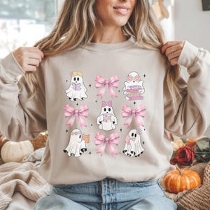 Puede incluir: Sudadera beige con un estampado de fantasmas de dibujos animados y lazos rosas. Los fantasmas blancos leen libros. Los lazos son de color rosa claro. El diseño tiene una estética caprichosa y linda, ideal para un look casual.