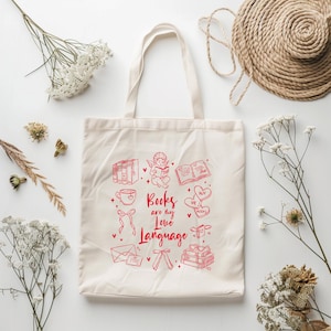 Puede incluir: Bolso tote de color crema con ilustraciones y texto en rojo. El diseño incluye libros, corazones, un cupido y la frase "Books are my Love Language". El bolso tiene dos asas y está hecho de una tela resistente.