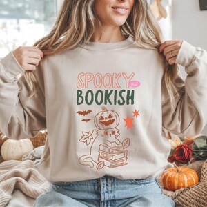 Puede incluir: Sudadera beige con el texto "SPOOKY but BOOKISH" en rosa y verde. Un gato con una calabaza en la cabeza está sentado sobre una pila de libros. Murciélagos, hojas y estrellas también están presentes en el diseño.