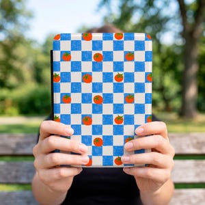 May include: A rectangular e-reader case featuring a repeating pattern of small red tomatoes with green stems centered within alternating blue and white checkered squares. The design has a textured appearance across the entire surface of the cover.