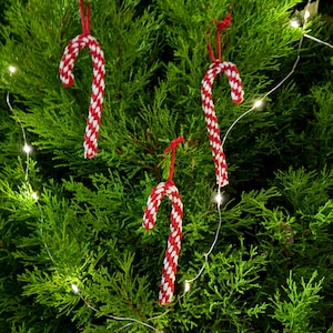 Könnte beinhalten: Drei rot-weiß gestreifte Zuckerstangen-Ornamente hängen an einem grünen Nadelbaum. Die Zuckerstangen bestehen aus gewebtem Material und sind an roten Schnüren aufgehängt. Eine Lichterkette mit kleinen weißen Lichtern ist um den Baum gewickelt.