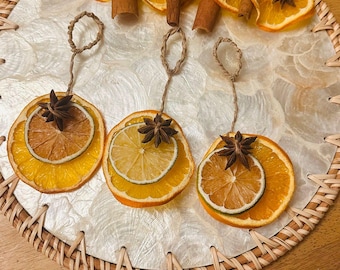 May include: A decorative arrangement featuring dried orange and lime slices, star anise, and cinnamon sticks. The items are displayed on a woven placemat with a textured white surface. Three ornaments hang from twine loops.