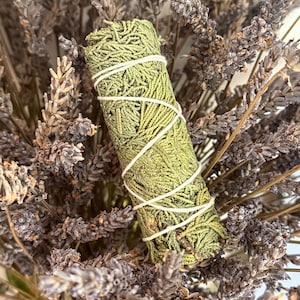 Puede incluir: Un manojo cilíndrico de hojas de cedro verde secas, envuelto firmemente con un hilo de algodón blanco fino, colocado verticalmente sobre un fondo denso y texturizado de ramas de lavanda morada seca con tallos y pequeños capullos visibles.