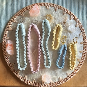 Op de afbeelding: Een verzameling handgemaakte macramé sleutelhangers in pastelkleuren, waaronder lichtblauw, roze en geel. Elke sleutelhanger heeft een unieke bedel, zoals een ster, bloem, kat of halve maan. De sleutelhangers worden gepresenteerd op een ronde geweven tray met decoratieve kristallen.