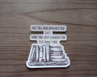 Peut inclure: Un autocollant avec le texte "NOT ALL WHO WANDER ARE LOST. SOME ARE JUST LOOKING FOR THE BOOKSTORE." L'autocollant représente une pile de livres avec un design d'étagère. L'autocollant est blanc avec du texte et des illustrations noirs.