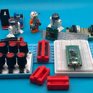 May include: A collection of electronic components and Lego figures, including a green circuit board on a breadboard, red components, and Star Wars Lego characters, all arranged on a blue surface.