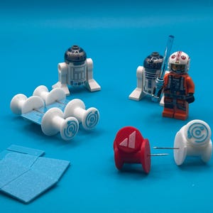 May include: A collection of Star Wars-themed Lego figures and accessories on a blue background. Includes two R2-D2 figures, a pilot figure with a lightsaber, and various small, white and red decorative pieces. The scene is brightly lit.