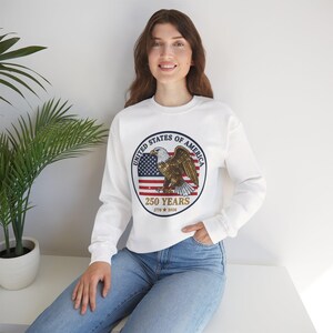 Puede incluir: Sudadera blanca con un diseño gráfico circular. El diseño incluye una bandera estadounidense, un águila calva y el texto "UNITED STATES OF AMERICA" y "250 YEARS 1776 2026". La sudadera tiene mangas largas.