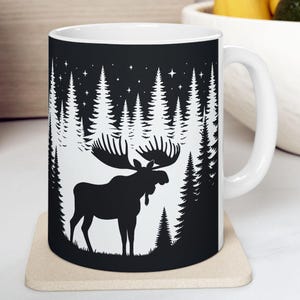 May include: White ceramic mug featuring a black silhouette of a moose in a forest scene. The design includes tall pine trees and a starry night sky. The mug has a curved handle and is set on a beige coaster.