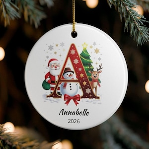 May include: A round white ceramic Christmas ornament featuring a large red letter A decorated with snowflakes. The design includes Santa Claus, a snowman, a green Christmas tree, and a reindeer. The name Annabelle and the year 2026 are printed below.