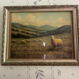 May include: Framed pastoral artwork featuring a flock of sheep grazing in a field with rolling hills in the background. The artwork includes a quote from Jeremiah 33:3. The frame is gold-toned.