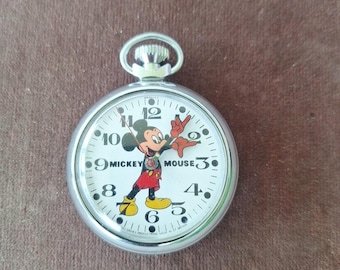 May include: A silver pocket watch with a white face featuring Mickey Mouse. The watch has black numerals and hands, with the Mickey Mouse character in the center. The watch is round and has a loop at the top.