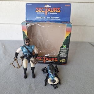 May include: A vintage Sectaurs action figure set featuring Mantor and Raplor. The Mantor figure is in a blue and white outfit, holding a bow and shield. The set includes a box with the Sectaurs logo and the text "Warriors of Symbion".