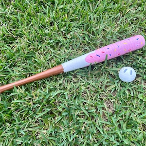 May include: A pink and white baseball bat with a donut design and a white baseball on a bed of green grass. The bat is approximately 28 inches long.