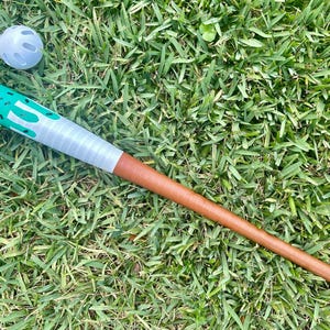May include: A baseball bat with a green and white design, a gray grip, and a brown handle lies on green grass next to a white baseball. The bat has a unique design and is made for playing baseball.