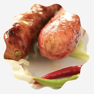 May include: Close-up of two cooked sausages with a glossy, reddish-brown exterior. The sausages rest on a bed of white cabbage and are accompanied by red and green chili peppers, suggesting a spicy dish.