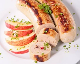 May include: Two browned sausages on a white plate, one whole and one sliced into rounds, accompanied by four thin red apple slices. The sausages and apple slices are garnished with small green parsley flakes and peppercorns.