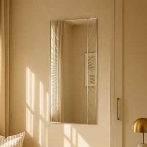 May include: A rectangular mirror with a silver frame and a sunburst design. The mirror is mounted on a beige wall. The room has a neutral color palette, with a beige curtain and a white cabinet. A gold lamp and a small picture frame are on a side table.