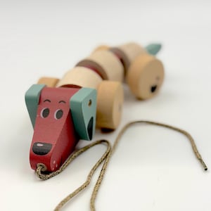 Può includere: Un giocattolo di legno a forma di cane da trascinare, con testa rossa, orecchie azzurre e corpo marrone. Il giocattolo ha ruote e un cordino per tirare. Il muso del cane ha occhi neri e naso nero. Il giocattolo è in legno e dipinto in vari colori.