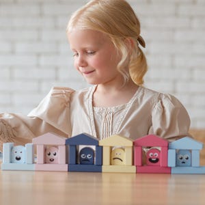Puede incluir: Un niño juega con un juego de bloques de madera de colores. Los bloques tienen forma de edificios y presentan diferentes expresiones faciales. Los bloques están pintados en colores pastel, incluyendo azul, rosa y amarillo. El niño sonríe y mira los bloques.