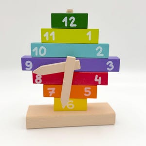 May include: A colorful wooden clock toy with a light wood base. The clock has stacked blocks in various colors, each labeled with a number from 1 to 12. The clock hands are made of light wood.